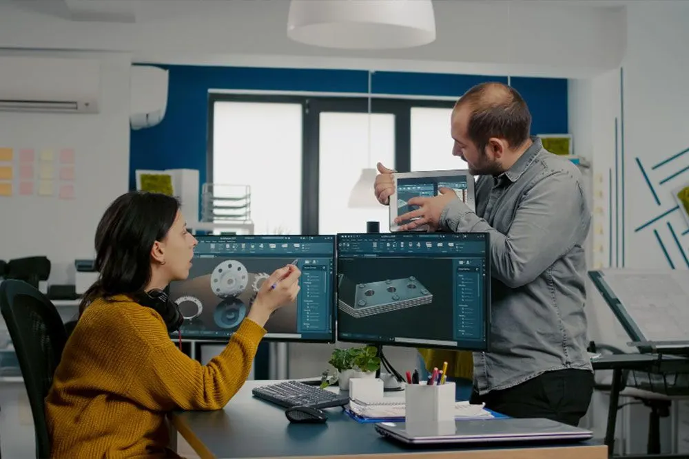 Two engineers reviewing 3D CAD models of machine parts on screens and a tablet